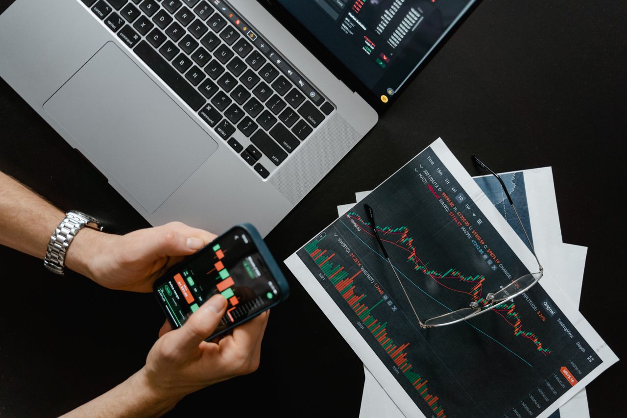 digital-01 Hands using smartphone beside laptop with stock charts, showcasing digital trading.